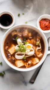 Top-view image of a bowl of homemade Hot and Sour Soup served on a white marble counter. The rich, savory broth is packed with mushrooms, tofu, bamboo shoots, and delicate egg ribbons. Garnished with fresh green onions and a drizzle of chili oil, this spicy and tangy soup is the perfect comfort food for any occasion.