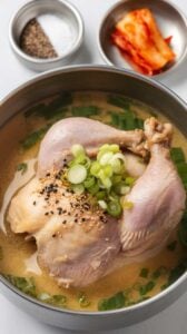 Top-down image of a bowl of Samgyetang (Korean Ginseng Chicken Soup) served on a white marble counter. The tender whole chicken sits in a light golden broth, infused with ginseng, jujubes, and garlic. Garnished with sliced green onions and served with a side of salt and black pepper, this nutritious dish is a Korean favorite for boosting energy and wellness.