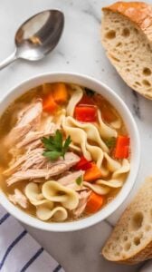 Top-down image of a bowl of Instant Pot Chicken Noodle Soup served on a white marble counter. The comforting soup is filled with shredded chicken, egg noodles, carrots, and celery in a flavorful broth. Garnished with fresh parsley and served with crusty bread for the perfect cozy meal.