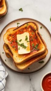Top-down image of crispy Air Fryer Texas Toast served on a plate. The thick slices are golden brown, buttery, and topped with garlic and parsley. Served with marinara sauce on the side for a delicious and easy garlic bread.