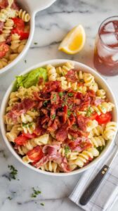 A photo of a creamy BLT pasta salad in a white bowl. The salad is filled with pasta, bacon, lettuce, and tomatoes. There's extra crumbled bacon and chopped parsley on top. A lemon wedge and a cold drink are placed beside the bowl. The salad is served on a marble kitchen counter, with a napkin and a fork beside it.
