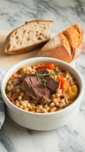 A photo of a steaming bowl of beef and barley stew served in a white ceramic bowl on a marble countertop. The thick, hearty stew is packed with tender beef, chewy barley, and colorful vegetables, garnished with a sprig of fresh thyme. A slice of crusty bread sits beside the bowl, ready for dipping.