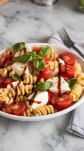 A photo of a white ceramic plate filled with Caprese pasta salad. The salad contains spiraled pasta, bright cherry tomatoes, creamy mozzarella, and ribbons of fresh basil. A drizzle of balsamic vinaigrette glistens on top. The plate sits on a marble kitchen counter, and there's a fork and napkin beside it. The background is blurred and contains a few kitchen items.