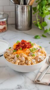 A photo of a white bowl filled with chicken bacon ranch pasta salad on a marble kitchen counter. The salad is garnished with extra bacon bits, shredded cheese, and chopped green onions. A spoon and a cloth napkin are placed beside the bowl. The background contains kitchen utensils and a plant.