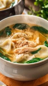 A bowl of chicken dumpling soup with fluffy dumplings, shredded chicken, carrots, celery, and fresh spinach in a golden broth, garnished with black pepper and parsley.
