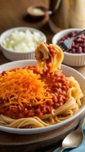 A serving of Cincinnati Chili over spaghetti, topped with shredded cheddar cheese, with diced onions and kidney beans on the side. The rich sauce coats the pasta for a classic Midwestern meal.