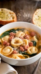 A bowl of hearty sausage and tortellini soup with sausage, tortellini, and spinach in a tomato broth, garnished with parmesan and black pepper, served with garlic bread.