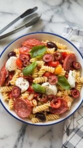 A photo of a serving bowl of Italian antipasto pasta salad on a marble counter. The salad is vibrant with visible chunks of salami, mozzarella, olives, and tomatoes. The pasta is garnished with fresh basil and cracked black pepper. There are tongs and a napkin beside the bowl.