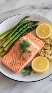 A photo of a plated serving of lemon herb salmon on a white ceramic plate, garnished with fresh dill and lemon slices. The salmon is served alongside garlic butter asparagus and a small portion of lemon quinoa. The background is a marble kitchen counter, adding a clean and elegant touch to the image.