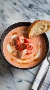 A photo of a deep bowl of creamy lobster bisque with a smooth, velvety texture. The bisque is garnished with chunks of tender lobster meat, a drizzle of cream, and a sprinkle of fresh chives. A slice of toasted baguette is placed on the side. The bowl is placed on a marble kitchen counter. There is a napkin and spoon nearby.
