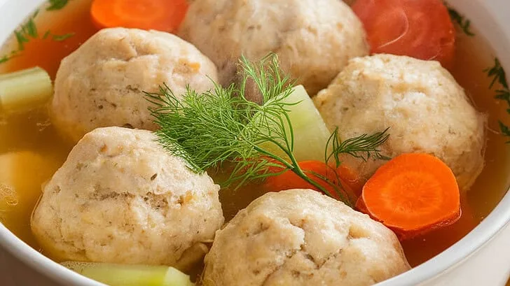 A bowl of matzo ball soup with a golden broth, fluffy matzo balls, carrots, celery, and fresh dill garnish. Served with warm challah bread for a complete meal.