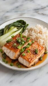 A photo of a plated serving of miso glazed cod with a caramelized, golden glaze. The cod is garnished with sesame seeds and chopped green onions. The dish is served alongside steamed jasmine rice and stir-fried bok choy. The presentation is on a white ceramic plate on a marble kitchen counter.