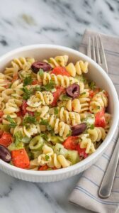 A photo of a white ceramic bowl filled with a zesty Italian pasta salad. The salad is garnished with chopped parsley and cracked black pepper. The salad features spiral pasta, vibrant vegetables, and olives, and is served chilled. The bowl rests on a marble kitchen counter, with a fork and a linen napkin beside it.