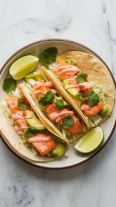 Top-down image of crispy Air Fryer Salmon Tacos served on a plate. The warm corn tortillas are filled with flaky salmon, crunchy cabbage, creamy avocado, and a drizzle of sauce. Garnished with fresh cilantro and lime wedges, these tacos are fresh and flavorful.