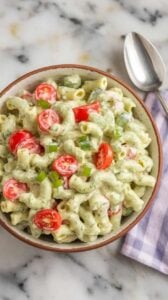 A photo of a ceramic bowl filled with avocado ranch pasta salad. The salad is creamy and colorful, with bright red tomatoes and green onions. The bowl is set on a marble kitchen counter. A spoon and a napkin are beside the bowl.