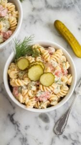 A photo of a white ceramic bowl containing a serving of dill pickle pasta salad on a marble kitchen counter. The salad is garnished with fresh dill and extra chopped pickles. There's a fork and a pickle spear next to the bowl for presentation.