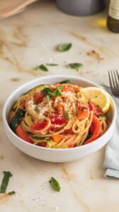 A photo of a white ceramic bowl filled with pasta primavera. The pasta is spaghetti tossed with sautéed vegetables, including bell peppers, zucchini, and carrots. The dish is topped with chopped basil and grated Parmesan cheese. A lemon wedge and cracked black pepper are sprinkled on top. The bowl sits on a marble kitchen counter, which is off-white. There is a fork and a napkin nearby. The background is blurred and contains a few kitchen items, such as a wooden spoon and a bottle of oil.