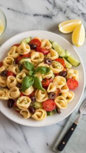 A photo of a white ceramic plate with a serving of tortellini pasta salad. The salad is made with cheese tortellini, vibrant vegetables such as cherry tomatoes, cucumber, and olives, and is tossed in a lemon dressing. The salad is topped with chopped basil leaves. There are lemon wedges on the side. The plate is placed on a marble counter. A fork and a napkin are also placed near the counter.