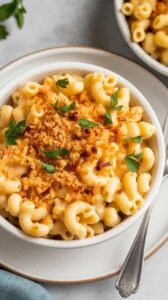A photo of a white ceramic plate containing a bowl of vegan mac and cheese. The mac and cheese is made with elbow pasta coated in a golden, creamy sauce. The dish is topped with toasted breadcrumbs and chopped parsley for extra flavor and texture. A fork is placed beside the dish. The background is clean and simple.