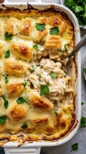 A close-up photograph of a creamy chicken casserole in a white ceramic baking dish. The dish contains golden-brown puff pastry pieces arranged in a lattice pattern on top, with a rich, creamy white sauce underneath. Fresh green parsley leaves are scattered across the surface as garnish. A silver serving spoon is inserted into the casserole, lifting up a portion that reveals shredded chicken and creamy sauce texture. The pastry pieces have a crispy, caramelized exterior with darker browned edges. The baking dish is positioned on a light gray textured surface, with fresh parsley leaves visible in the background. The lighting is bright and even, highlighting the glossy surface of the sauce and the golden color of the pastry. The image is shot from a top-down angle with the baking dish filling the entire frame.