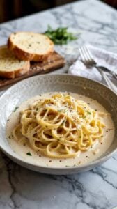 A high-quality food photograph of fettuccine alfredo in a white ceramic bowl with a textured pattern around the rim. The pasta is coated in a creamy white sauce and garnished with freshly grated Parmesan cheese and chopped parsley. In the background, there is a wooden cutting board with two slices of toasted bread. A white cloth napkin and silver fork are visible in the upper right corner. The entire scene is set on a white and gray marble surface with distinctive veining patterns. The lighting is soft and natural, creating subtle shadows that enhance the texture of the pasta and the marble surface. The composition is shot from a slightly elevated angle, with the pasta taking up approximately two-thirds of the frame. The depth of field is shallow, with the pasta in sharp focus while the background elements are slightly blurred.