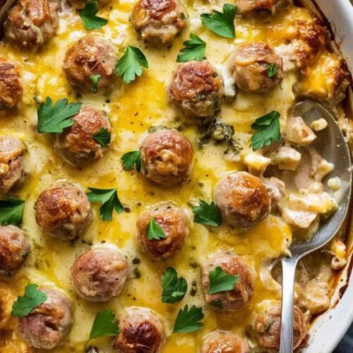 A close-up photograph of a white ceramic baking dish filled with a creamy casserole dish. The dish contains perfectly browned meatballs arranged in a grid pattern, topped with melted cheddar cheese that has a bright yellow color and glossy surface. Fresh green parsley leaves are scattered across the top as garnish. A silver serving spoon is partially visible on the right side of the dish. The casserole has a rich, creamy sauce visible between the meatballs. The baking dish has a decorative bubble pattern around its rim. The background shows a dark blue-gray napkin and fresh parsley sprigs slightly out of focus. The lighting is bright and even, highlighting the golden-brown crust of the meatballs and the melted cheese.