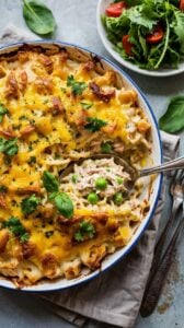 A high-quality food photography shot of a creamy tuna casserole in a white ceramic baking dish with a blue rim. The casserole has a golden-brown melted cheese topping with crispy, toasted breadcrumbs and bacon bits scattered throughout. Fresh green parsley and basil leaves are sprinkled on top for garnish. A silver serving spoon is lifting a portion of the casserole, revealing layers of creamy white sauce, tuna, and small green peas. In the background, there is a small white bowl containing a fresh green salad with red cherry tomatoes. The scene is set on a light gray textured surface with a folded beige linen napkin visible in the bottom right corner. The lighting is bright and natural, highlighting the glossy cheese surface and creating subtle shadows that add depth to the composition.