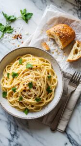A high-quality food photograph of spaghetti carbonara in a white ceramic bowl with a speckled gray rim, positioned on a white marble surface. The pasta is garnished with fresh green parsley leaves and black pepper, creating a creamy white sauce coating the noodles. To the right of the bowl is a torn piece of golden-brown artisanal bread on white parchment paper, with visible crumb texture. A silver fork rests on a light beige linen napkin to the right of the bowl. Fresh parsley sprigs are scattered on the left side of the marble surface. Red chili flakes are visible near the bread. The lighting is bright and natural, creating subtle shadows and highlighting the glossy texture of the pasta. The marble surface has distinctive gray veining patterns running through it. The composition is shot from a top-down angle with the bowl positioned slightly off-center.