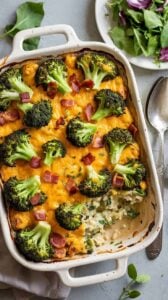 A high-quality food photograph of a creamy broccoli and bacon casserole in a white ceramic baking dish with handles. The casserole has a golden-brown melted cheese topping with fresh green broccoli florets and crispy bacon bits scattered throughout. A portion has been served, revealing a light-colored creamy rice base underneath the cheese topping. The baking dish is positioned on a light gray surface with a white napkin partially visible. In the background, there is a small white plate with fresh green salad leaves and purple cabbage. A silver spoon is placed on the right side of the frame. Fresh green basil leaves are scattered around the dish. The lighting is bright and even, creating a professional food photography aesthetic with soft shadows. The image is shot from a slightly elevated angle, showing the texture and layers of the dish clearly.