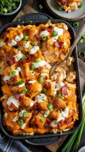 A top-down view of a loaded tater tot casserole in a black rectangular Staub baking dish. The casserole has a golden-brown melted cheese topping with bacon bits, sliced green onions, and dollops of sour cream arranged across the surface. The tater tots are golden brown and perfectly crispy, arranged in a uniform pattern. A silver serving spoon is positioned in the bottom right corner of the dish. In the background, there is a small white plate with a slice of the casserole, and a small bowl of fresh chopped green herbs. The dish is photographed on a dark wooden surface, with fresh green onions visible at the edges of the frame. The text "SOUDAY" and "ORUDANG" is visible on the black baking dish. The lighting is warm and professional, highlighting the glossy cheese and creating appetizing shadows across the dish.